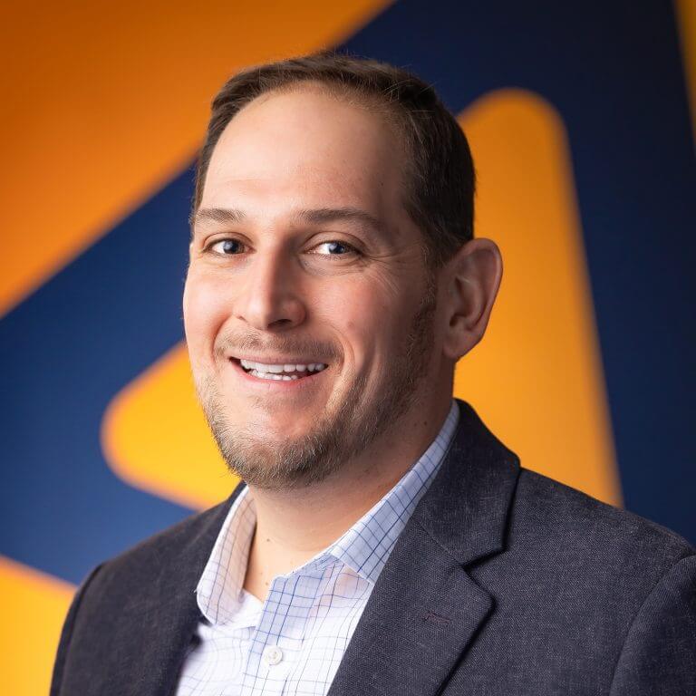 A many in gray suit, white checkered shirt, with glasses in front of blue and orange background.