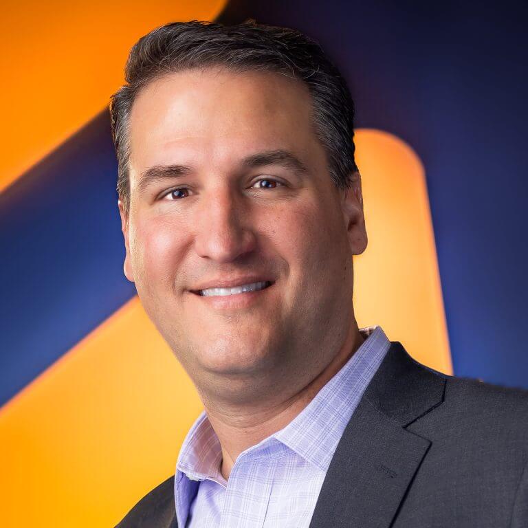 A many in gray suit, checkered purple shirt, in front of blue and orange background.