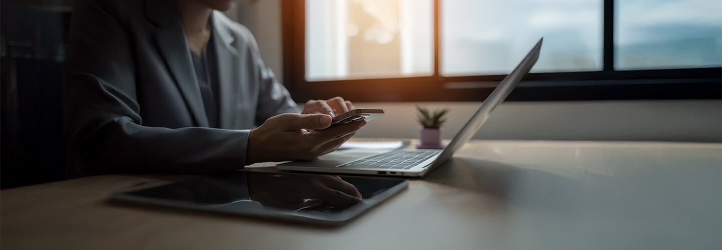 A person sitting at a desk with cellphone in hand in front of a laptop